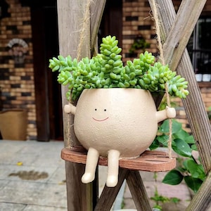 Może przedstawiać: Beżowa ceramiczna doniczka w kształcie osoby, z uśmiechniętą twarzą i ramionami, trzymająca drewnianą huśtawkę. Doniczka jest wypełniona zielonymi roślinami sukulentów. Huśtawka jest zawieszona na sznurze.