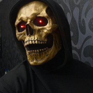 Puede incluir: Una máscara de calavera dorada con ojos rojos brillantes, combinada con una sudadera con capucha negra. La máscara presenta dientes detallados y una estructura ósea realista, ideal para Halloween.