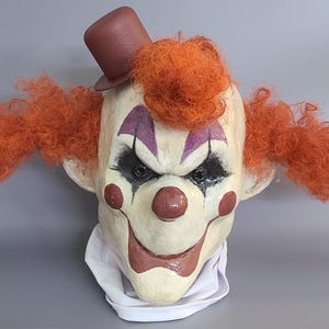 Puede incluir: Máscara de payaso aterrador con sombrero de copa marrón y pelo naranja. La máscara tiene una cara blanca con detalles rojos y morados, y ojos negros. Un cuello blanco es visible en la base de la máscara.