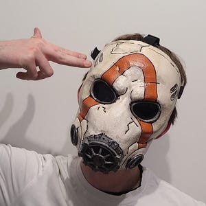 May include: A white and orange mask with black accents, featuring a distressed design. The mask has large eye openings and a series of circular vents. The mask is worn by a person wearing a white shirt.