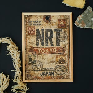 May include: A framed vintage-style print featuring the letters "NRT" above "TOKYO" and "JAPAN." The print includes postage stamp designs and the words "PAR AVION." The frame is made of light-colored wood.