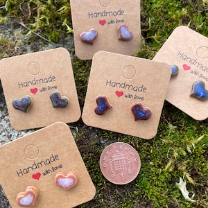 May include: A collection of handmade heart-shaped stud earrings in various colors, presented on brown cards. Each card is printed with "Handmade with love." A one penny coin is also visible. The earrings are on a mossy surface.