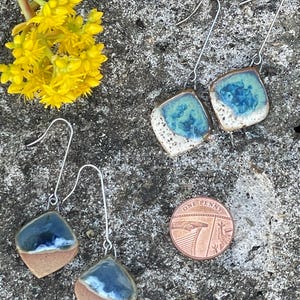 May include: Handmade ceramic earrings with a square shape and long silver hooks. The earrings feature a two-tone design with a brown base and a top section in shades of blue and white. A penny is next to the earrings.