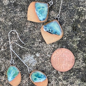 May include: Handmade ceramic earrings with a distinctive design. Each earring has a brown base with a turquoise and blue glazed top. The earrings have long, silver-coloured hooks. A one penny coin is included for size reference.