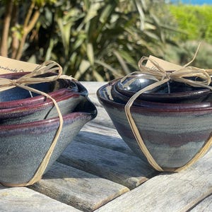 May include: A collection of handmade ceramic bowls in graduated sizes, tied with raffia. The bowls feature a dark blue-grey exterior and a reddish-brown rim. The bowls are presented on a wooden surface, with a tag that says "Handmade".