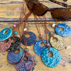 May include: A collection of circular ceramic pendants with various glazed textures and colours, including blue, green, and brown, suspended on thin brown leather cords. A pair of tortoise-shell patterned sunglasses rests above the pendants.