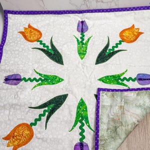 May include: A handmade quilt with a floral design of orange and purple tulips. The tulips have green stems and leaves, arranged in a circular pattern on a white background. The quilt is edged with a purple dotted fabric.