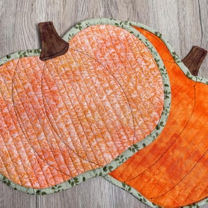 Peut inclure: Deux sets de table en forme de citrouille matelassées. Les citrouilles sont orange avec des tiges marron et des bordures vertes à motifs de feuilles. L'une a un motif, l'autre est orange uni. Sur une surface en bois.