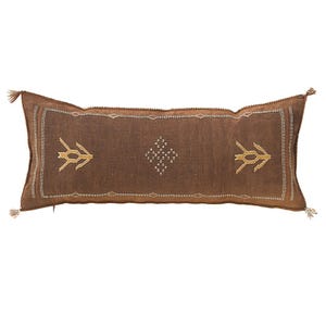 May include: A rectangular brown decorative pillow with embroidered geometric designs. The pillow has a central panel with a diamond pattern and two stylized arrow-like shapes. Tassels adorn each end. The pillow is designed for home decor.