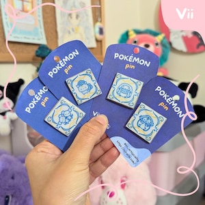 May include: Four square Pokémon pins on blue backing cards. Each pin features a different Pokémon character in a blue and yellow tile design. The cards are labeled "Pokémon pin" and include a website address.