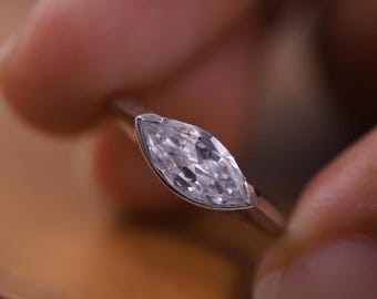 May include: A close-up of a marquise-cut diamond ring. The diamond is set in a thin, silver band. The ring is held by a person's fingers, with a wooden surface in the background. The diamond sparkles with many facets.