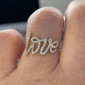 May include: A gold ring spelling "love" in a cursive font. The letters are set with small, sparkling gemstones. The ring is a piece of jewellery. The design is elegant and eye-catching, suitable for everyday wear or special occasions.