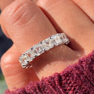 Peut inclure: Gros plan d'une bague d'éternité en diamants. La bague est ornée d'une série de diamants de taille carrée sertis dans un anneau en métal blanc. La bague est portée sur un doigt, avec une manche bordeaux en arrière-plan.