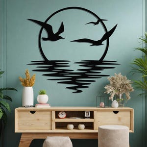 Puede incluir: Arte mural de metal negro que representa gaviotas volando frente a un sol circular sobre el agua. La obra de arte está montada en una pared turquesa sobre un mueble de madera con objetos decorativos y plantas. El diseño crea una escena serena inspirada en la naturaleza.