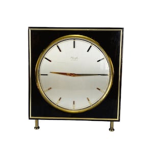 May include: A vintage square clock with a black wooden frame and a gold-colored circular clock face. The clock face has a silver background with black and gold accents, and the clock reads "Ringele Automatic". The clock is supported by two small gold-colored legs.