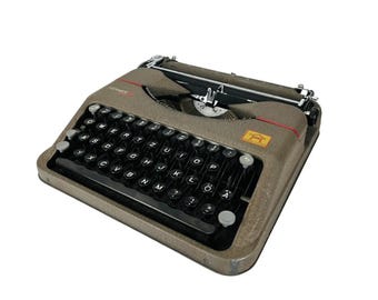 May include: A vintage Hermes 3000 typewriter in a muted brown color. The typewriter features a black keyboard with white letter keys and a red accent line. The Hermes logo is visible on the right side.