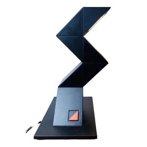 Könnte beinhalten: Eine moderne schwarze Schreibtischlampe mit geometrischem Design. Die Lampe hat einen zickzackförmigen Arm und einen rechteckigen Sockel. Ein orangefarbenes Dreieck ist auf der Vorderseite sichtbar. Die Lampe ist für eine gezielte Beleuchtung konzipiert.