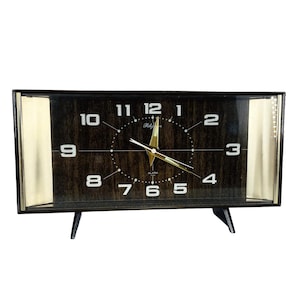 May include: A vintage rectangular alarm clock with a dark wood grain face and gold accents. The clock face features white numerals, gold hands, and the word "Rhythm". The clock has a black frame and legs.