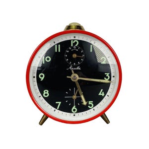 Puede incluir: Reloj despertador Mauthe vintage rojo y negro. La esfera redonda negra muestra números blancos y manecillas doradas. El reloj, de unos 10 cm de diámetro, está enmarcado en rojo y descansa sobre patas doradas.