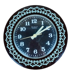 May include: A round Weimar quartz clock with a dark brown frame and a black face. The clock has white numerals and hands, with a light blue decorative border. The text "WEIMAR" and "QUARTZ" are visible on the clock face.