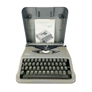 Puede incluir: Una máquina de escribir Hermes Baby gris vintage con su estuche a juego. El teclado tiene teclas negras con letras blancas. El estuche está abierto, revelando un manual con una foto de la máquina de escribir y la palabra "Baby".