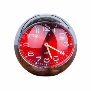 May include: A vintage Kaiser alarm clock with a red face and white numerals. The clock has a chrome and wood-grain finish. The hands are cream-colored. The clock is a sphere shape.