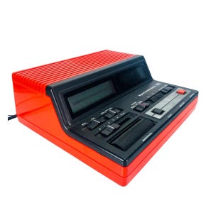 May include: A retro red and black Synchrotec 200 radio alarm clock. The clock features a digital display, various buttons, and a slider. The top section is red with horizontal ridges. The front panel is black.