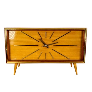 May include: A mid-century modern clock with a light wood face and a dark wood frame. The clock has gold-colored hands and markers, and is supported by four tapered wooden legs. The clock measures approximately 18 inches wide.