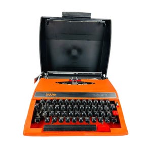 Puede incluir: Máquina de escribir Brother Deluxe 650TR naranja con teclado y carcasa negros. La máquina de escribir tiene teclas negras y detalles en rojo. La carcasa está abierta, revelando los componentes internos. El logotipo de Brother es visible.