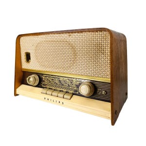 Puede incluir: Radio Philips vintage con una carcasa de madera y una cubierta de altavoz tejida. La radio presenta perillas y botones de color crema, un borde dorado y el nombre de la marca "PHILIPS" impreso en la parte delantera.
