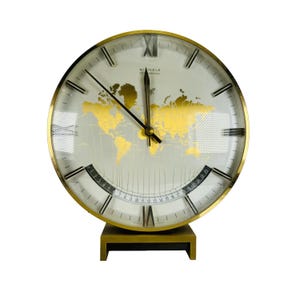 Könnte beinhalten: Eine goldfarbene Kinzle International Uhr mit Weltkarten-Design auf dem Zifferblatt. Die Uhr hat schwarze Zeiger, römische Ziffern und eine Datumsanzeige. Die Uhr steht auf einem rechteckigen Sockel.