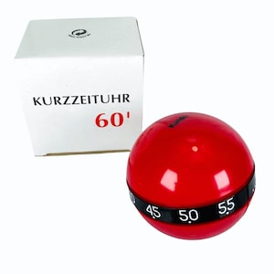 Puede incluir: Un temporizador de cocina esférico rojo con una banda negra que muestra números. El temporizador está junto a una caja blanca con el texto "KURZZEITUHR 60'".