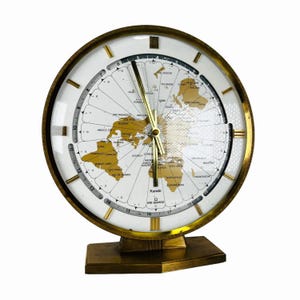 Könnte beinhalten: Eine Vintage-Weltzeituhr mit einem goldfarbenen Metallrahmen und -fuß. Das Zifferblatt hat einen weißen Hintergrund mit einer goldenen Weltkarte und schwarzen Ziffern. Die Zeiger der Uhr sind goldfarben. Der Markenname "Kundo" ist sichtbar.