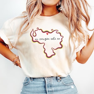 May include: An ivory short-sleeve t-shirt featuring a graphic outline of the map of Venezuela with a yellow, blue, and red border. Inside the map, the text reads "mi corazón está en" in black cursive script with a small red heart above it.