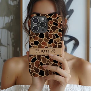 May include: A phone case with a giraffe print design in shades of brown, tan, and black. The case has a tan band with the name "KATE" printed on it. The phone case is held in front of a person.