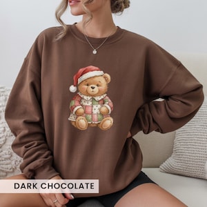 May include: Dark chocolate-colored sweatshirt featuring a watercolor illustration of a teddy bear wearing a Santa hat and a patchwork coat. The bear is centered on the front of the sweatshirt. The text "DARK CHOCOLATE" is at the bottom.