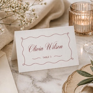 Puede incluir: Una tarjeta de sitio color crema con el nombre "Olivia Wilson" en escritura burdeos, enmarcada por un borde decorativo. Las palabras "TABLE 3" están debajo del nombre. La tarjeta está colocada sobre una superficie de mármol, con una vela y una copa de vino en el fondo.