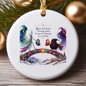 May include: A round white ceramic ornament featuring a colorful illustration of two whimsical figures meeting on a bridge above a snowy village. The text reads: Some Christmas meetings quietly become the beginning of everything. It has a gold hanging cord.