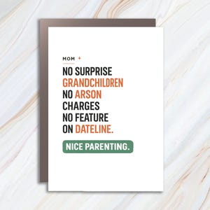 May include: A white greeting card with a brown border. The card reads "MOM + NO SURPRISE GRANDCHILDREN NO ARSON CHARGES NO FEATURE ON DATELINE. NICE PARENTING." in a mix of black, orange, and green text.