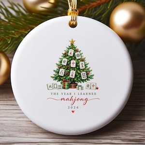 May include: A round white ceramic ornament featuring a green Christmas tree decorated with Mahjong tiles. Below the tree, the text reads THE YEAR I LEARNED mahjong 2026 with a small red heart. The ornament hangs from a gold cord.