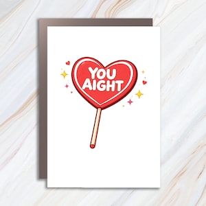 May include: A white greeting card featuring a red heart-shaped lollipop illustration in the center. The candy has the white text YOU AIGHT printed on it. Small yellow four-pointed stars and tiny red hearts surround the lollipop on the white background.