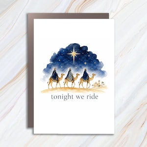 May include: A white greeting card featuring a watercolor illustration of three figures riding camels across a desert landscape toward a distant building. Above them is a dark blue starry sky with a large gold star. The text reads tonight we ride.