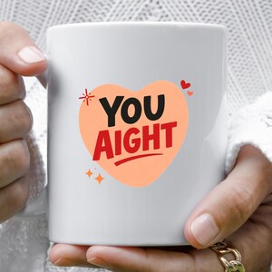 May include: White ceramic mug with a peach-colored heart design. The heart features the words "YOU AIGHT" in black and red text. Small red hearts and starburst graphics accent the design. The mug is held by a person wearing gold rings.