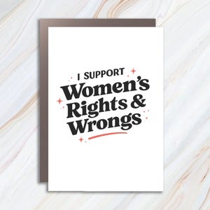 May include: A white greeting card featuring the black text I SUPPORT Women's Rights & Wrongs. The design includes small coral-colored star accents and a horizontal line beneath the text, presented on a dark gray backing card.