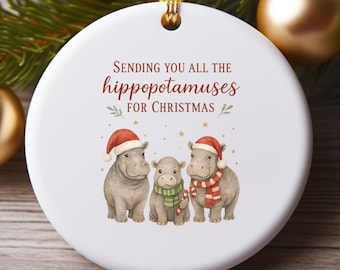 May include: A white ceramic Christmas ornament with a gold hanging cord. The ornament features three hippopotamus illustrations wearing Santa hats and scarves. Text reads "Sending you all the hippopotamuses for Christmas."