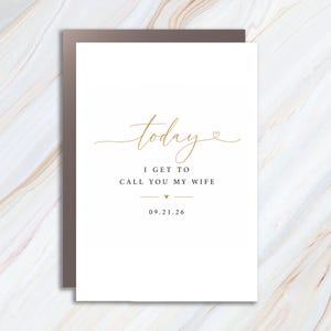 May include: A white rectangular greeting card with gold script text that reads today followed by black text that reads I GET TO CALL YOU MY WIFE and the date 09.21.26. The design includes two small gold hearts and horizontal lines.