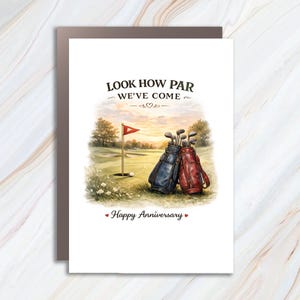Könnte beinhalten: Eine Grußkarte mit einer Golfplatz-Illustration, die eine blaue und eine rote Golftasche, eine Fahne und einen Golfball zeigt. Der Text lautet LOOK HOW PAR WE'VE COME und Happy Anniversary, verziert mit kleinen Herzen und einem Herzsymbol.