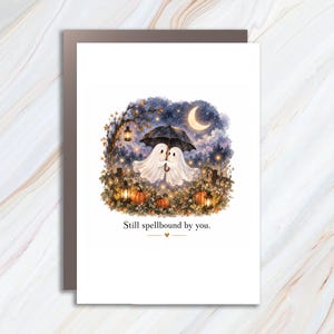 May include: A white greeting card featuring a watercolor illustration of two ghosts under a black umbrella, surrounded by orange pumpkins, glowing lanterns, and a crescent moon in a night sky, with the text Still spellbound by you.