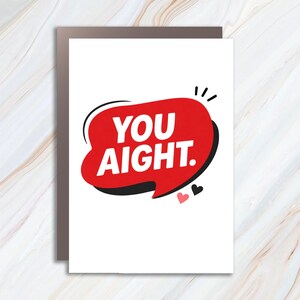 May include: A white greeting card featuring a red speech bubble with a black drop shadow. The text YOU AIGHT. is printed in bold white capital letters inside the bubble. A small pink heart and a small black heart are positioned below the bubble.