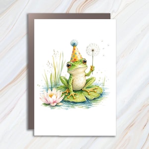 May include: A watercolor illustration of a green frog wearing a yellow party hat with colorful polka dots. The frog sits on a large green lily pad in water, holding a white dandelion seed head. A pink water lily and tall green reeds are nearby.